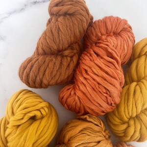 May include: A collection of yarn skeins in various shades of orange, yellow, and brown. The yarn is chunky and soft, perfect for knitting or crocheting.
