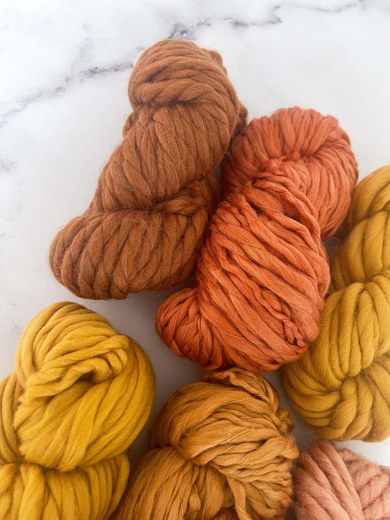 May include: A collection of yarn skeins in various shades of orange, yellow, and brown. The yarn is chunky and soft, perfect for knitting or crocheting.