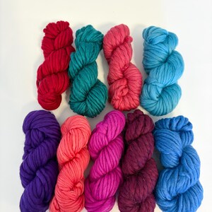 May include: Ten skeins of colorful, chunky yarn in a variety of shades. The colors include red, teal, pink, light blue, purple, coral, magenta, burgundy, and dark blue. The yarn is arranged in two rows of five skeins each, against a white background.