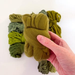 May include: A hand holding a bundle of olive green wool roving, with other bundles of green wool in varying shades in the background. The wool is soft and fluffy, ideal for fiber art projects.