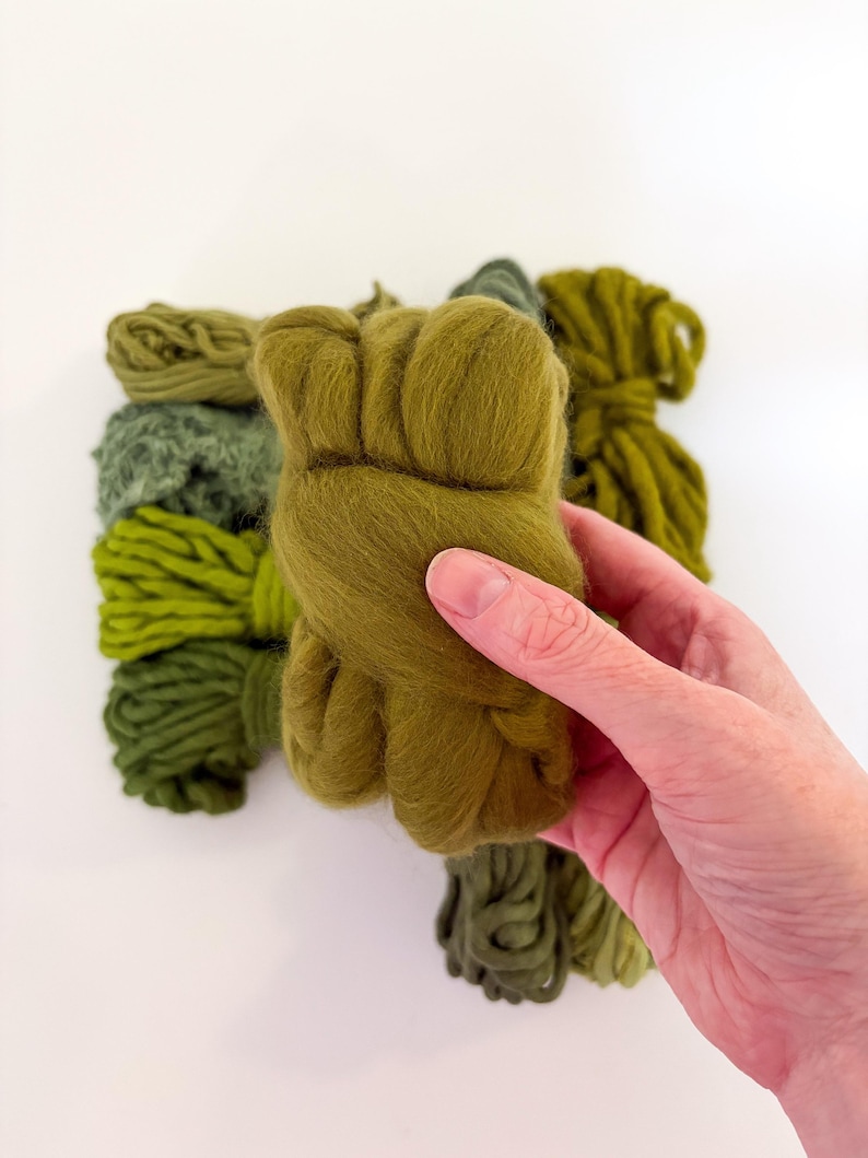 May include: A hand holding a bundle of olive green wool roving, with other bundles of green wool in varying shades in the background. The wool is soft and fluffy, ideal for fiber art projects.