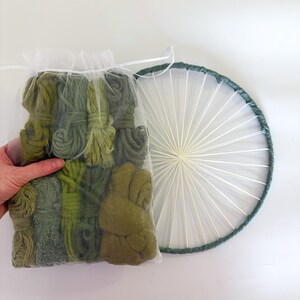 May include: A clear bag filled with various shades of green yarn bundles, held by a hand. Next to it is a circular weaving hoop with a dark green border and white string spokes, creating a sunburst design.