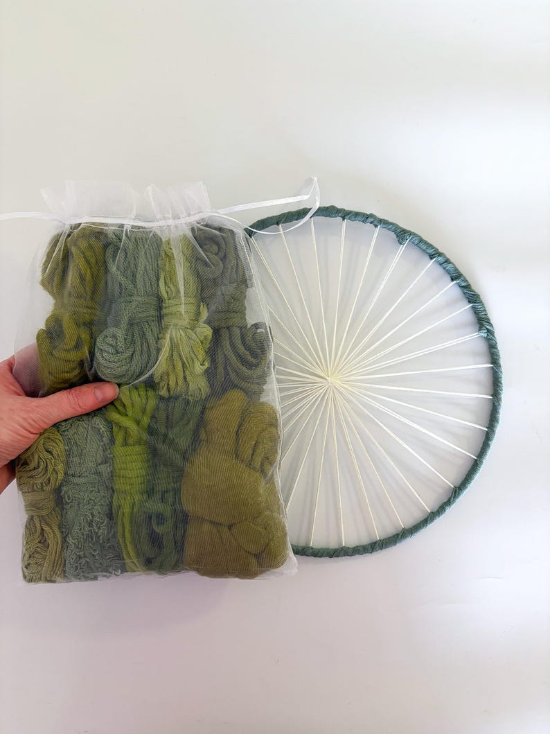 May include: A clear bag filled with various shades of green yarn bundles, held by a hand. Next to it is a circular weaving hoop with a dark green border and white string spokes, creating a sunburst design.