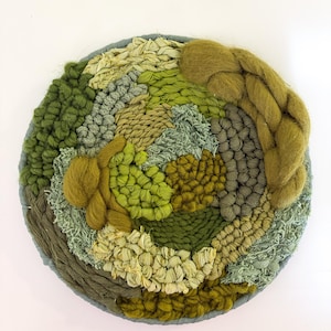 May include: A round, woven wall hanging with a variety of green and tan textures. The piece features a circular design with a gray border. The textures include braided yarn, looped yarn, and woven fabric, creating a tactile and visually interesting piece.