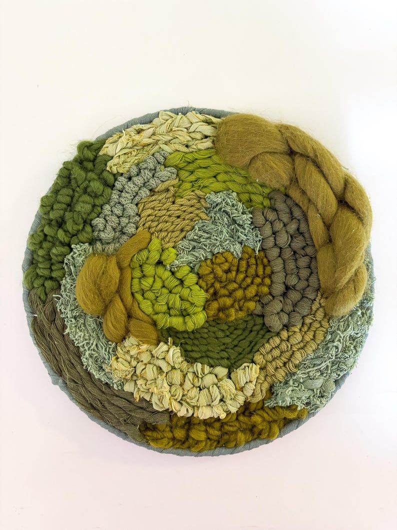 May include: A round, woven wall hanging with a variety of green and tan textures. The piece features a circular design with a gray border. The textures include braided yarn, looped yarn, and woven fabric, creating a tactile and visually interesting piece.