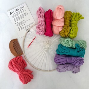 May include: A craft kit for making a wall hanging with colorful yarn. The kit includes a wooden hoop, a comb, a needle, and 10 skeins of yarn in various colors, including pink, green, purple, and blue. The kit is labeled "Lush Little Loops Candy Rainbow Weaving Kit".