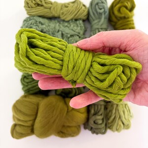 May include: A hand holding a bundle of bright green yarn, with several other bundles of green yarn in varying shades of green in the background. The yarn appears to be soft and textured, suitable for knitting or other crafts.