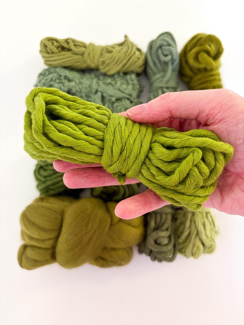 May include: A hand holding a bundle of bright green yarn, with several other bundles of green yarn in varying shades of green in the background. The yarn appears to be soft and textured, suitable for knitting or other crafts.