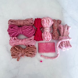 May include: Assortment of pink and red yarn and fabric scraps in various textures and shades. The yarn is arranged in rows, with the fabric scrap in the centre.