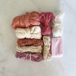 May include: A collection of pink, white, and beige yarn, string, and fabric. The items are arranged in a row on a white marble surface.