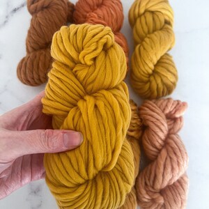 May include: Five skeins of chunky yarn in shades of brown, orange, and yellow. The yarn is arranged in a pile, with the yellow skein on top.