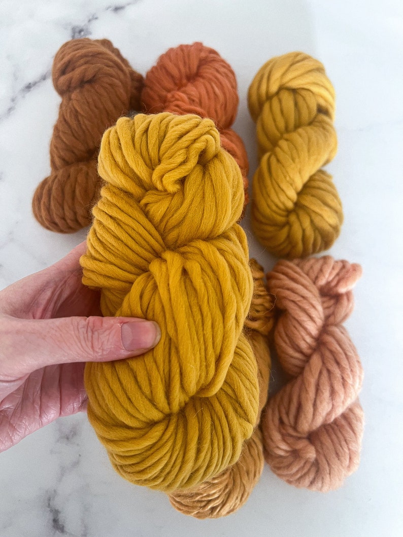 May include: Five skeins of chunky yarn in shades of brown, orange, and yellow. The yarn is arranged in a pile, with the yellow skein on top.