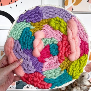 May include: A colorful circular wall hanging made with yarn in shades of pink, purple, green, blue, and yellow. The yarn is woven into a variety of textures and patterns, creating a unique and eye-catching design.