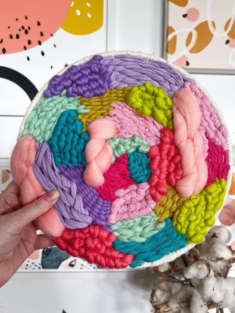 May include: A colorful circular wall hanging made with yarn in shades of pink, purple, green, blue, and yellow. The yarn is woven into a variety of textures and patterns, creating a unique and eye-catching design.