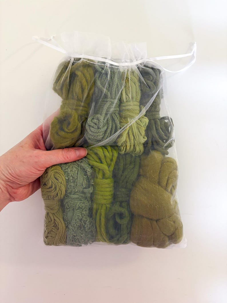 May include: A clear, drawstring bag filled with various shades of green yarn. The yarn is bundled and tied, showcasing a range of textures and hues, from light olive to deep forest green. The bag is held by a hand against a white background.