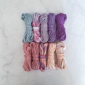 May include: Eight spools of cotton cord in various shades of pink, purple, and blue. The cords are arranged in a row, with the lightest colour at the top and the darkest colour at the bottom.