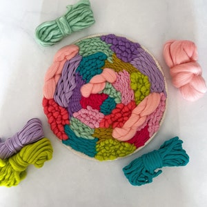 May include: A colorful yarn wall hanging with a variety of colors including pink, green, purple, and yellow. The yarn is woven into a circular pattern and is displayed on a white background.