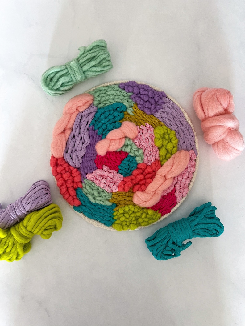 May include: A colorful yarn wall hanging with a variety of colors including pink, green, purple, and yellow. The yarn is woven into a circular pattern and is displayed on a white background.