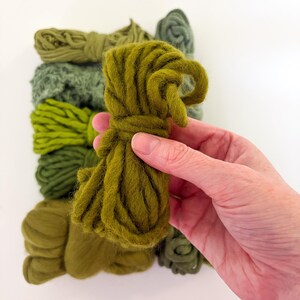 May include: A hand holds a bundle of olive green yarn, with several other bundles of varying shades of green yarn in the background. The yarn appears to be thick and textured, suitable for crafting projects.