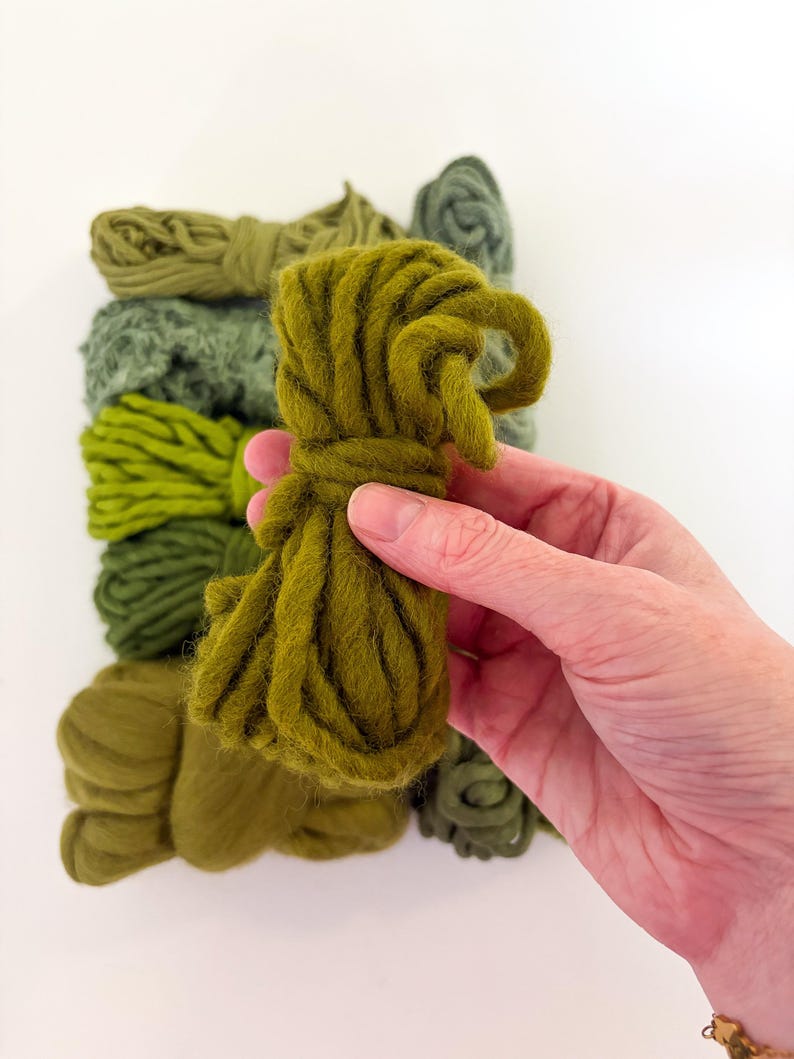 May include: A hand holds a bundle of olive green yarn, with several other bundles of varying shades of green yarn in the background. The yarn appears to be thick and textured, suitable for crafting projects.