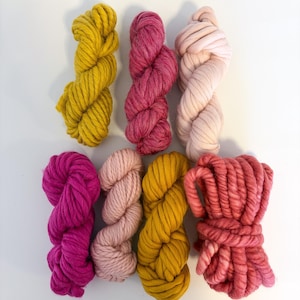 May include: A collection of eight skeins of chunky yarn in various shades of pink, yellow, and peach. The yarn is arranged in a neat, organized display, showcasing the different colors and textures. The yarn appears to be soft and inviting.