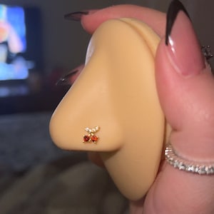 May include: A gold nose stud featuring a cherry design. The cherry has two red gems and a cluster of clear gems above. The nose stud is on a light skin tone.