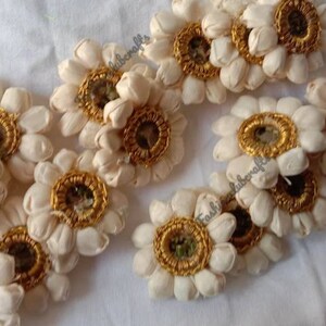 K&ouml;nnte beinhalten: Eine Sammlung von 12 wei&szlig;en Stoffblumenverzierungen mit goldenen Verzierungen und einem kleinen Spiegel in der Mitte. Die Blumen sind in einem zuf&auml;lligen Muster auf einer wei&szlig;en Oberfl&auml;che angeordnet.