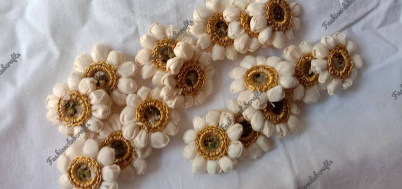 K&ouml;nnte beinhalten: Eine Sammlung von 12 wei&szlig;en Stoffblumenverzierungen mit goldenen Verzierungen und einem kleinen Spiegel in der Mitte. Die Blumen sind in einem zuf&auml;lligen Muster auf einer wei&szlig;en Oberfl&auml;che angeordnet.