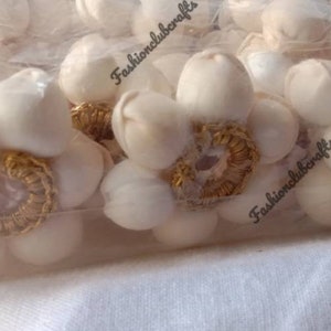 K&ouml;nnte beinhalten: Nahaufnahme einer T&uuml;te mit wei&szlig;en Stoffblumenverzierungen mit goldenen Verzierungen. Die Verzierungen befinden sich in einer durchsichtigen Plastikt&uuml;te mit dem Aufdruck "Fashionclubcrafts" auf der T&uuml;te.