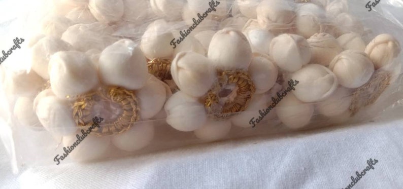 K&ouml;nnte beinhalten: Nahaufnahme einer T&uuml;te mit wei&szlig;en Stoffblumenverzierungen mit goldenen Verzierungen. Die Verzierungen befinden sich in einer durchsichtigen Plastikt&uuml;te mit dem Aufdruck "Fashionclubcrafts" auf der T&uuml;te.