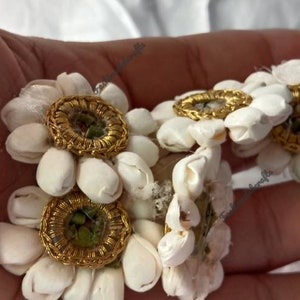 K&ouml;nnte beinhalten: F&uuml;nf wei&szlig;e Stoffblumenverzierungen mit goldenen Verzierungen und einem kleinen gr&uuml;nen Stein in der Mitte. Die Blumen sind in einer Gruppe angeordnet und werden in einer Hand gehalten.