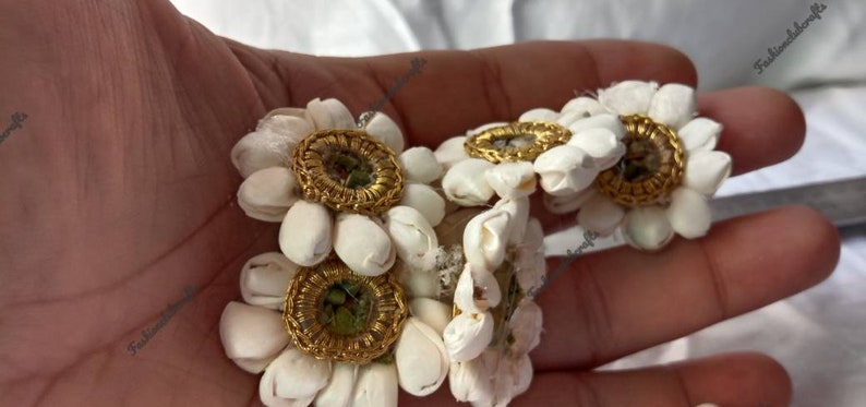 K&ouml;nnte beinhalten: F&uuml;nf wei&szlig;e Stoffblumenverzierungen mit goldenen Verzierungen und einem kleinen gr&uuml;nen Stein in der Mitte. Die Blumen sind in einer Gruppe angeordnet und werden in einer Hand gehalten.