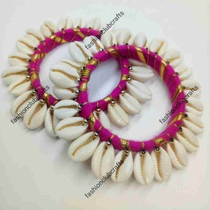 May include: Two pink and gold fabric bracelets with white shell beads. The bracelets are made with a pink fabric base and gold thread accents. The shells are arranged in a circle around the fabric.