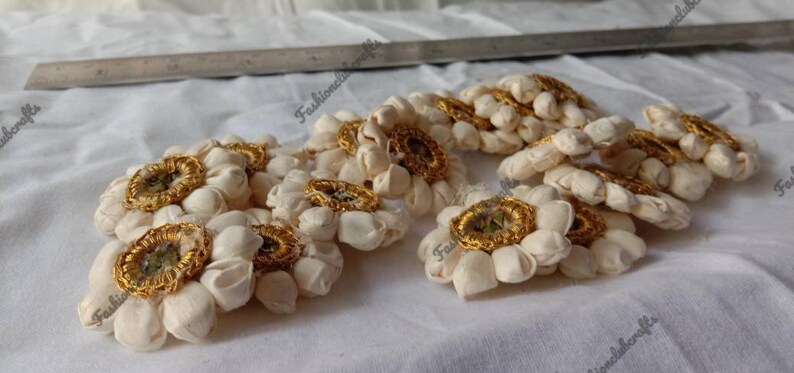 K&ouml;nnte beinhalten: Eine Gruppe von 10 wei&szlig;en Stoffblumenverzierungen mit goldenen Zentren. Die Blumen sind in einer Gruppe auf einer wei&szlig;en Stoffoberfl&auml;che angeordnet.