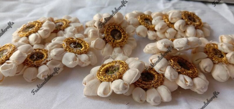 K&ouml;nnte beinhalten: Ein Set mit 8 wei&szlig;en Stoffblumen-Verzierungen mit goldenen Verzierungen. Jede Blume hat einen Mittelkreis mit goldener Umrandung und einem gr&uuml;nen Zentrum.