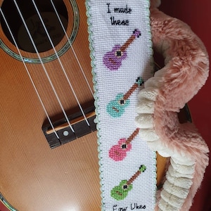 May include: A white bookmark with a green border and a cross-stitch design of four ukuleles in different colors. The bookmark reads "I made these Four Ukes".