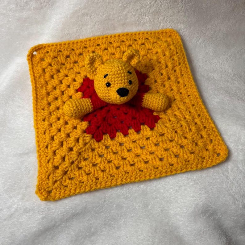 May include: A handmade yellow crochet baby blanket featuring a Winnie the Pooh character. The Pooh bear is yellow with a red shirt and black eyes. The blanket is square-shaped and has a textured pattern.