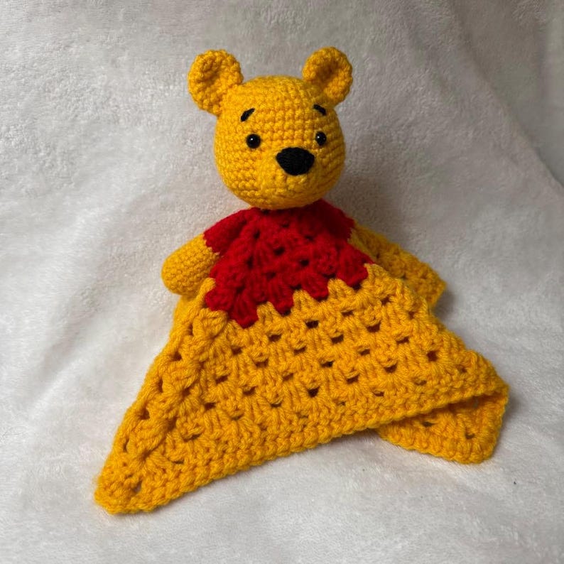 May include: A handmade yellow crochet bear with a red bib and a yellow blanket. The bear has a black nose and small black eyes. The blanket is a square shape with a textured pattern.