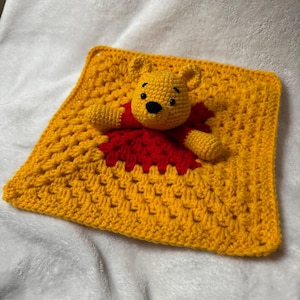 May include: A handmade crochet baby lovey featuring a yellow Winnie the Pooh character on a yellow square blanket. The bear has a black nose and is wearing a red shirt. The blanket has a textured pattern.