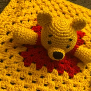 May include: A handmade crochet Winnie the Pooh bear toy with a yellow body and a red shirt. The bear is lying on a yellow and red crochet blanket. The toy has black eyes and a black nose.
