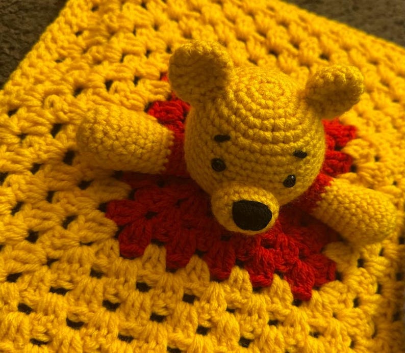 May include: A handmade crochet Winnie the Pooh bear toy with a yellow body and a red shirt. The bear is lying on a yellow and red crochet blanket. The toy has black eyes and a black nose.