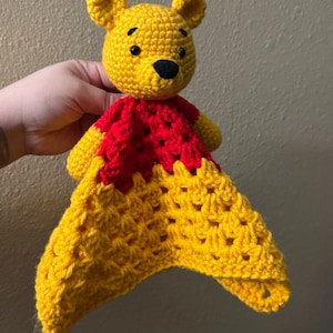 May include: A handmade crochet Winnie the Pooh lovey in yellow and red. The bear's head is yellow with black eyes and nose. The body is red, and the blanket portion is yellow with a textured pattern. A soft, cuddly item for babies and children.