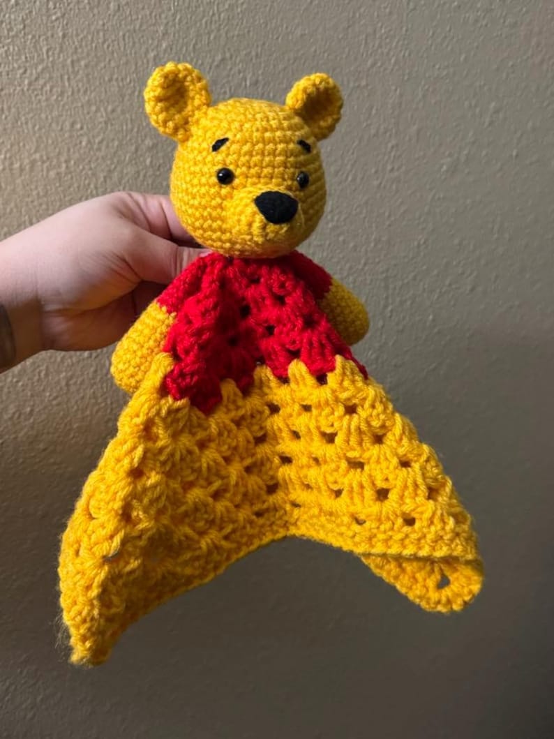 May include: A handmade crochet Winnie the Pooh lovey in yellow and red. The bear's head is yellow with black eyes and nose. The body is red, and the blanket portion is yellow with a textured pattern. A soft, cuddly item for babies and children.