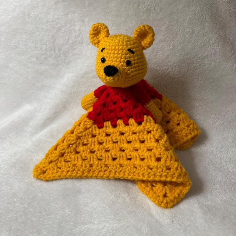 May include: Handmade crochet baby lovey featuring a yellow bear head with black eyes and nose, wearing a red shirt. The bear is attached to a yellow, open-weave blanket. A soft, cuddly toy for babies.