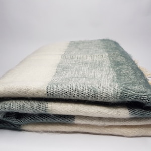 Op de afbeelding: Gevouwen deken met een kleurblokontwerp in crème, grijs en blauwgroen. De deken heeft een zachte, getextureerde uitstraling en een franje langs één rand. De deken is netjes opgevouwen en toont het kleurenpatroon.