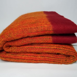 May include: Folded orange and red blanket. The blanket is made of a textured material with horizontal stripes. The top edge is a solid deep red color, while the rest of the blanket is a mix of orange and red hues.