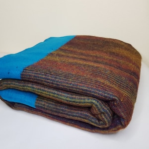 May include: Folded blanket with a vibrant color scheme. The blanket features a striking combination of teal blue and earthy tones, including shades of brown, orange, and yellow. The texture appears to be woven, with visible horizontal stripes.