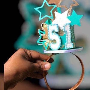 May include: A birthday headband with a teal, white, and gold color scheme. The headband features the number "51" and star decorations. The headband is held by a person.