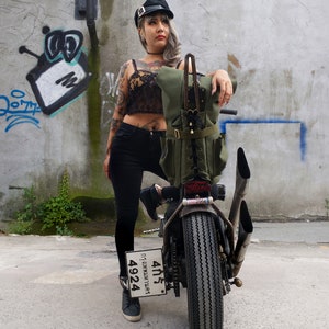 Op de afbeelding: Een groene canvas motorrugzak met meerdere zakken en leren accenten is gemonteerd op een aangepaste motorfiets. De motorfiets heeft een zwarte band, chromen uitlaatpijpen en een kentekenplaat. De achtergrond is een verweerde betonnen muur met graffiti.
