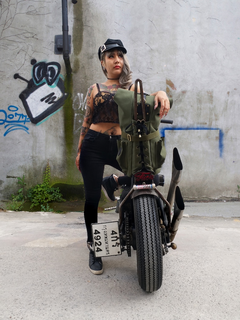 Op de afbeelding: Een groene canvas motorrugzak met meerdere zakken en leren accenten is gemonteerd op een aangepaste motorfiets. De motorfiets heeft een zwarte band, chromen uitlaatpijpen en een kentekenplaat. De achtergrond is een verweerde betonnen muur met graffiti.